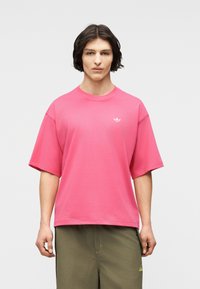 Άτομο που φοράει φωτεινό ροζ T-shirt Adidas με μικρό λευκό λογότυπο στο στήθος και ελιά πράσινο παντελόνι, σε απλό φόντο.