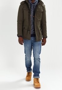 Veste matelassée vert olive avec fermetures éclair et poches, sweat à capuche gris en dessous, jean bleu et bottes beige avec œillets métalliques et lacets.
