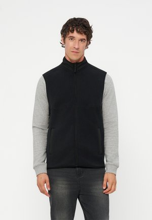 Homme portant un gilet noir zippé par-dessus une chemise à manches longues gris clair, debout devant un fond blanc uni.