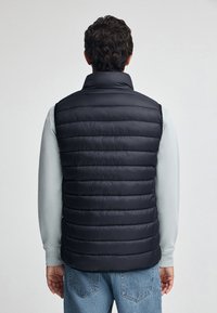 Czarna kamizelka puchowa z wysokim kołnierzem, pikowanym wzorem i gładką fakturą. Nosi się ją na jasnoszarej bluzie z długim rękawem oraz niebieskich dżinsach.