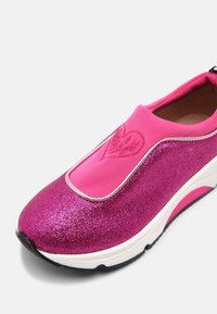 Zapatillas sin cordones en fucsia con un exterior brillante, detalle de corazón en relieve, cuello elástico rosa y suela blanca con acentos rosas.