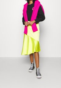 Sort langærmet skjorte med et cirkulært logo, lys pink strikket sweater draperet over skuldrene, og en satin gul asymmetrisk nederdel. Sorte sneakers.