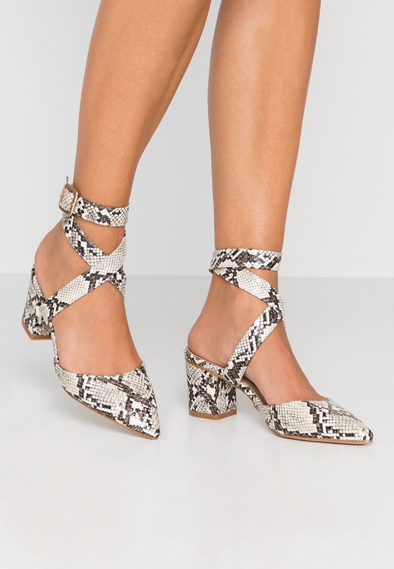 Sandales à talons imprimées serpent avec brides de cheville, dotées d'un bout pointu et d'un talon carré. Fabriquées en matériau synthétique dans des tons de blanc et de noir.