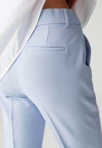 Helle blaue maßgeschneiderte Hose mit glatter Textur, die eine Gesäßtasche und ein schlichtes Bunddesign aufweist, teilweise von einem weißen Hemd bedeckt.