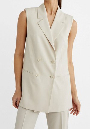 Blazer beige sans manches à double boutonnage avec revers porté sur un pantalon beige assorti, visible de la nuque jusqu'au milieu de la cuisse.