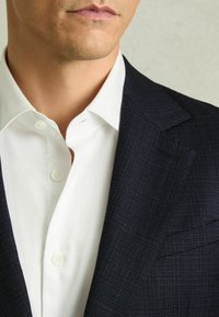 Tume mere blazer, millel on peen kontrollmustus, koos valge särgiga, millel on ümara kaelusega ja nööbid. Sile kangast tekstuur.