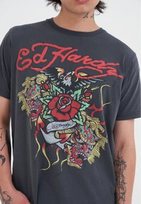 Camiseta negra con un gráfico detallado de un dragón, una rosa y un águila, con colores vibrantes y el texto "Ed Hardy" en rojo.