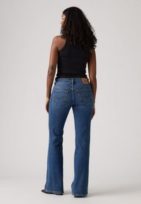 Jean en denim bleu évasé avec une taille mi-haute, présentant une étiquette en cuir et des accents de couture sur les poches arrière.