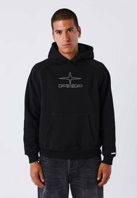 Jonge man met kort haar, gekleed in een zwarte hoodie met een witte ster en tekstontwerp, gecombineerd met donker gewassen jeans, staand voor een effen achtergrond.