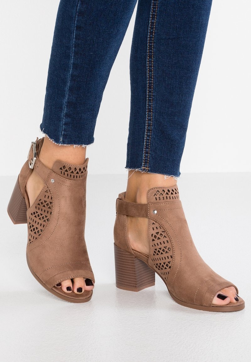 Bottines ouvertes en daim marron avec motifs ajourés, talons carrés, brides à la cheville, portées avec un jean bleu foncé à ourlet effiloché.