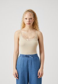 Šviesiai ruda (beige) liemenėlė su subtiliu nėriniu aplink iškirpę, derinama su laisvais mėlynais džinsais, turinčiais elastinę juostą ir raištelį.