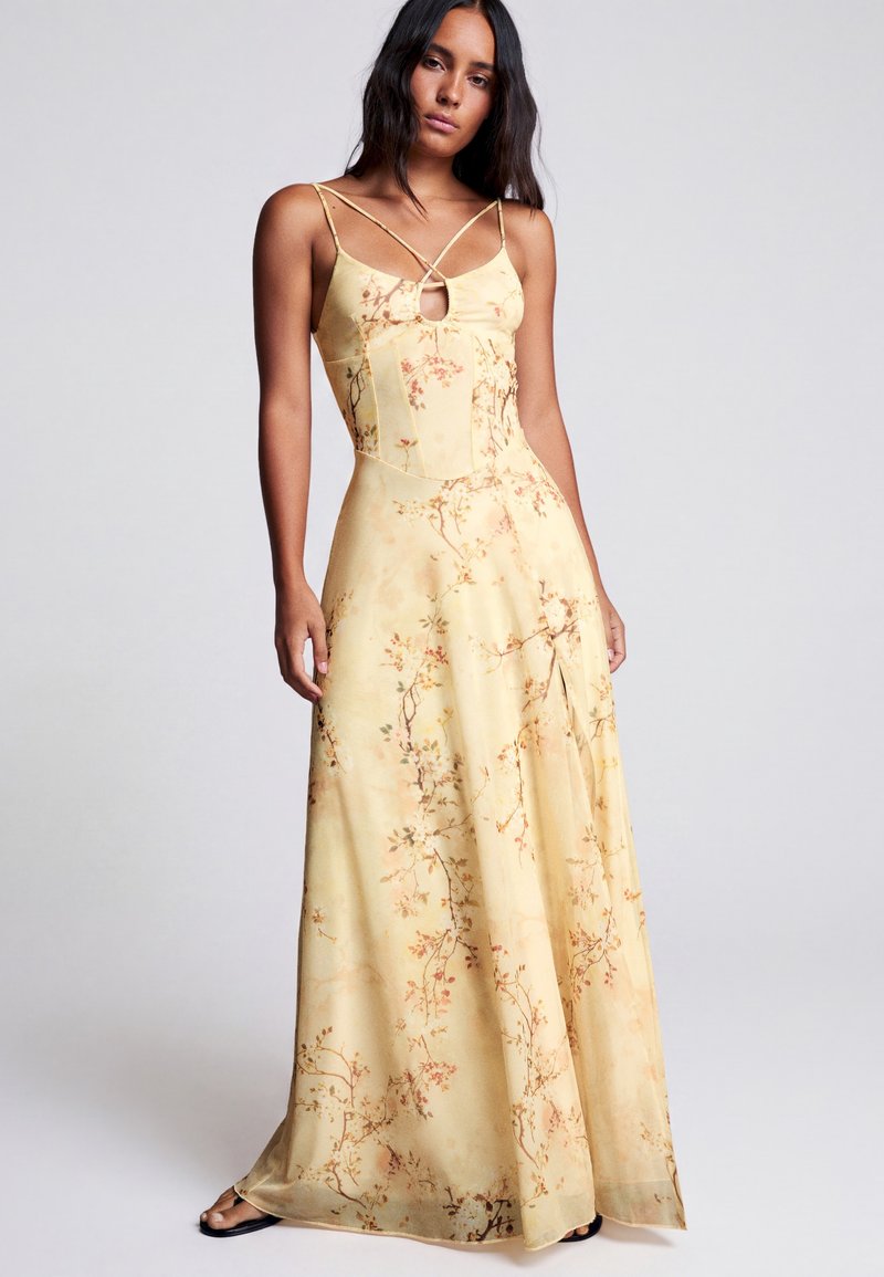 Femme portant une robe maxi jaune à fleurs avec de fines bretelles et un petit détail œillet à l'avant, debout devant un fond clair uni.