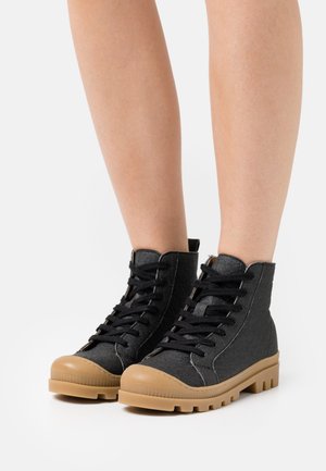 Bottes noires à lacets jusqu'à la cheville avec semelles en caoutchouc couleur tan, portées sur des jambes nues, posées sur un fond blanc.