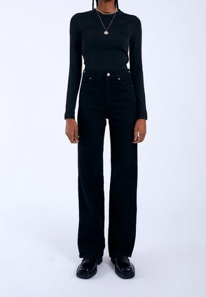Person iført højtaljede sorte jeans, sort langærmet tætsiddende top, sorte sko og en halskæde med en rund vedhæng, stående på hvid baggrund.