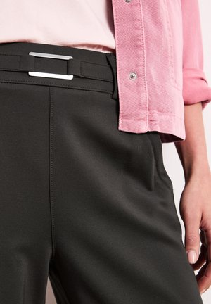 Persoon die zwarte broek draagt met een zwarte riem en een roze shirt met lange mouwen opgestroopt, hand rustend langs het lichaam.