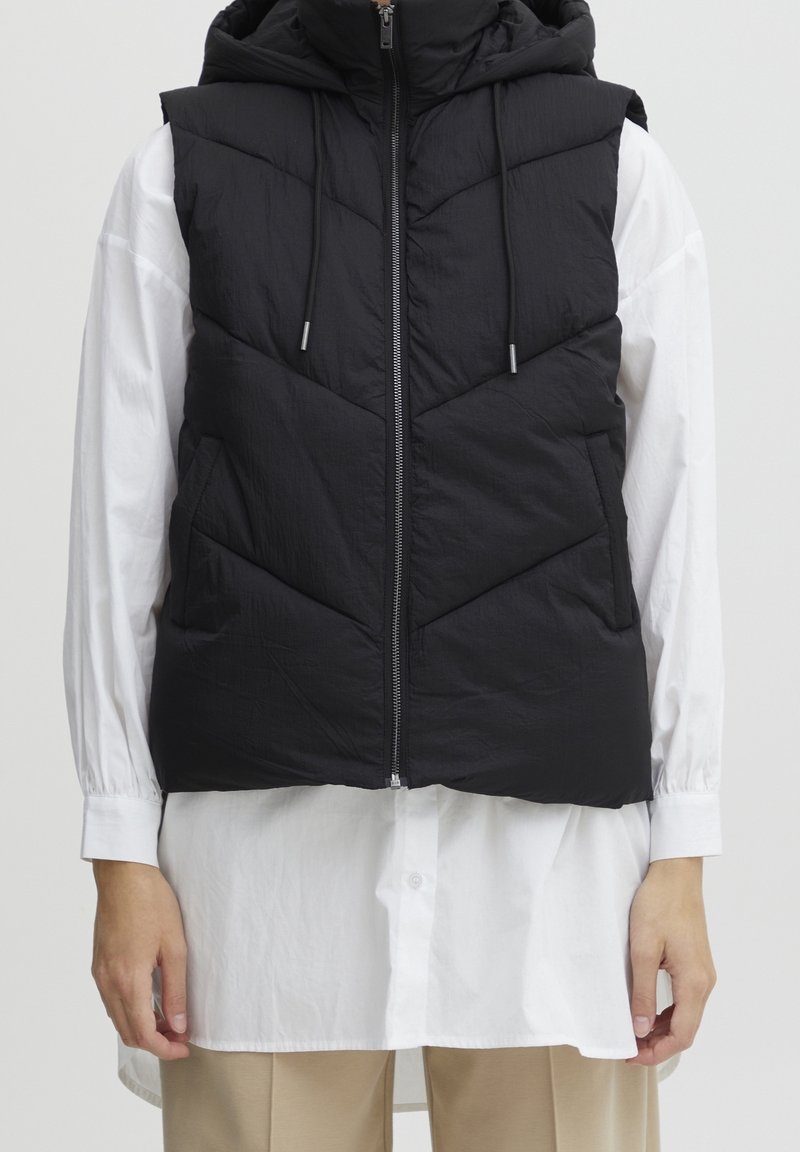 Personne portant un gilet noir matelassé sans manches avec capuche par-dessus une longue chemise blanche boutonnée et un pantalon beige, debout devant un fond uni.