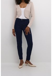 Beige kurzer Cardigan über einem weißen Oberteil, kombiniert mit dunkelblauen Skinny-Jeans und hellbeigen, spitzen High Heels.
