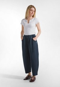 Kobieta ubrana w biały krótki top, luźne ciemnoniebieskie spodnie z kieszeniami oraz ciemnobrązowe buty na sznurowadła, stojąca z rękami w kieszeniach.