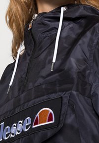 Svart nylonjacka med dragkedja i halsen, vita snören och en patch-logotyp. Har en slät yta och en delvis synlig huva.