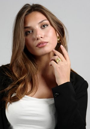 Goldfarbener Blumenring und passende Ohrringe, getragen mit einem schwarzen Blazer über einem weißen Oberteil. Sanft gewelltes braunes Haar umrahmt das Gesicht. Grauer Hintergrund.