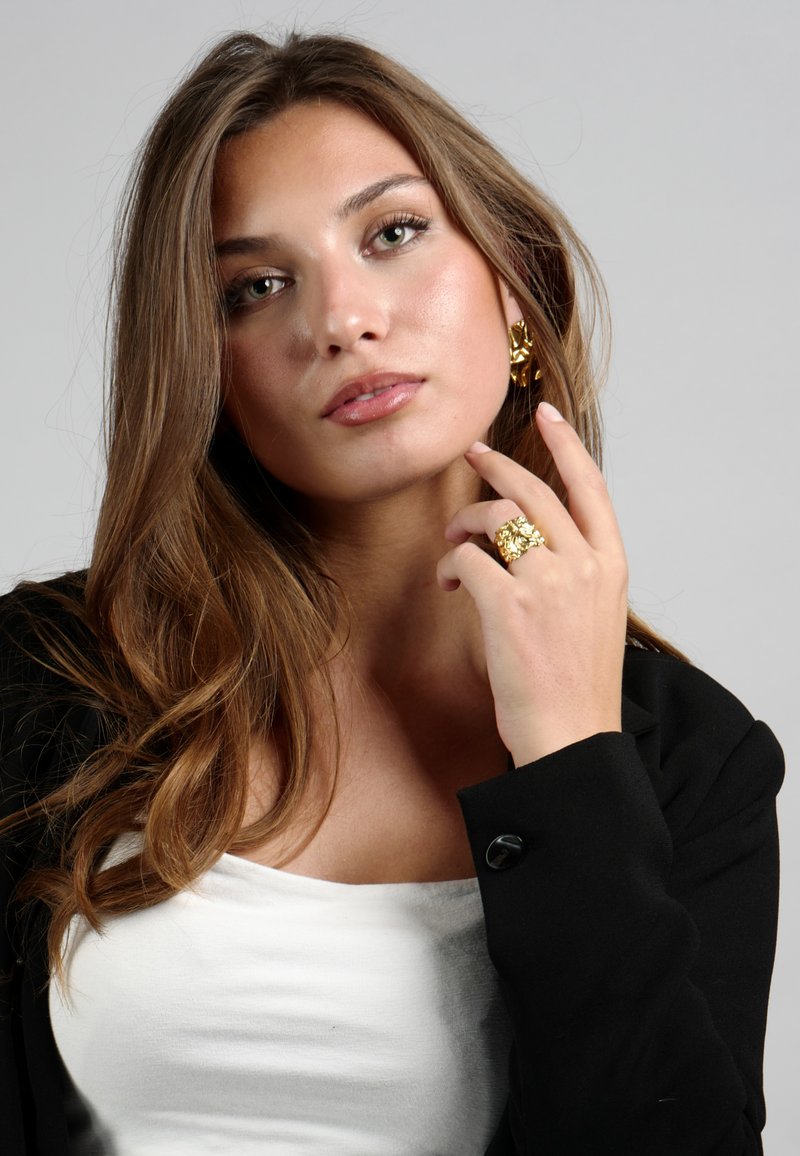 Anillo floral de oro y pendientes a juego, llevados con un blazer negro sobre una blusa blanca. Cabello castaño suave y ondulado enmarca el rostro. Fondo gris.