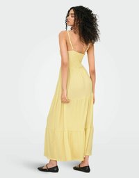 Robe maxi jaune avec des fines bretelles, un corsage smocké et une jupe en plusieurs couches. Fabriquée en tissu léger avec une texture douce. Des ballerines noires sont visibles.