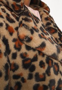 Leopardmönstrad fuskpälsjacka med en mjuk textur, överdimensionerad krage och framträdande svarta och orange fläckar på en beige bakgrund.