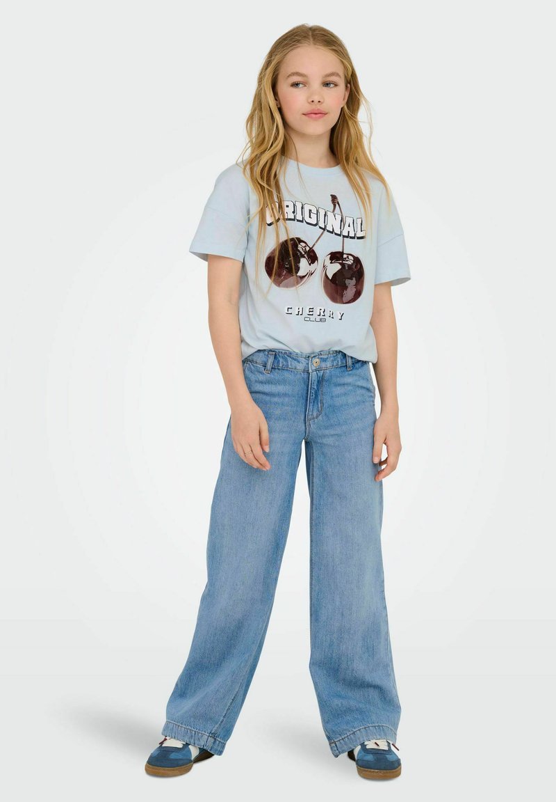 Jasnoniebieska grafika na koszulce z motywem wiśni oraz napisem, która jest zestawiona z jasnoniebieskimi spodniami o szerokich nogawkach. Ubranie uzupełniają niebieskie buty z białymi akcentami.