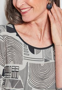 Schwarz-weiß gemusterte Bluse mit abstrakten Designs und geschwungenen Linien. Das Modell trägt runde, gestreifte Ohrringe und präsentiert einen runden Ausschnitt.