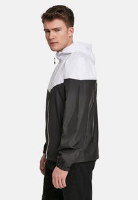 Veste légère pour homme en noir et blanc, avec fermeture éclair à l'avant, capuche ajustable et poignets élastiques, dotée d'une finition lisse et brillante.