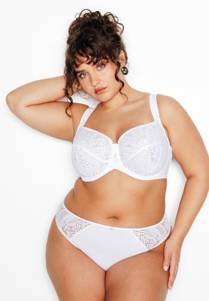 Femme aux courbes généreuses avec des cheveux bouclés portant un soutien-gorge en dentelle blanche et une culotte assortie, posant avec un bras levé sur un fond blanc.