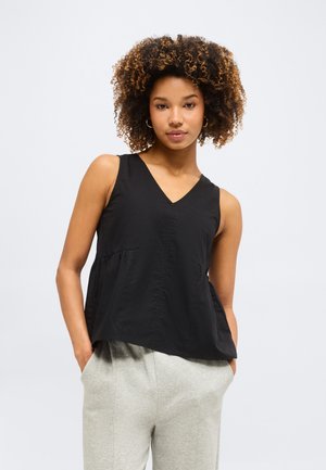 Młoda kobieta o kręconych włosach nosi bezrękawnik z czarnego materiału z dekoltem w serek oraz jasnoszare spodnie, stoi z rękami w kieszeniach na jednolitym tle.