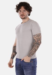 Camiseta de manga corta gris claro con un diseño texturizado, cuello redondo y forma ajustada. Combinada con jeans azules y con tatuajes visibles.