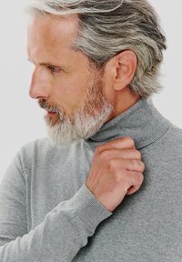 Szary sweter z golfem wykonany z miękkiego materiału, z prążkowanym kołnierzem i mankietami. Model delikatnie trzyma kołnierz, z jasnoszarymi włosami i brodą.