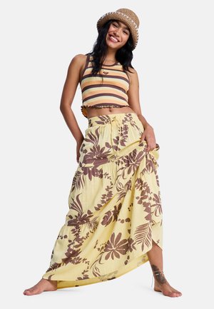 Smiling woman barefoot wearing a striped crop top, a wide yellow floral skirt, a woven hat, and ankle bracelets, posing against white background.