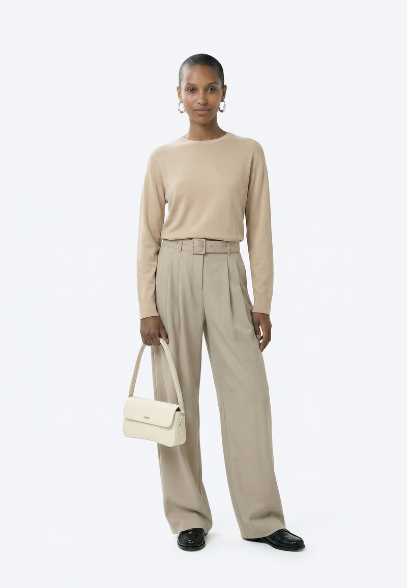 Donna con capelli corti in piedi, indossa una maglia beige a maniche lunghe, pantaloni larghi a vita alta con cintura, scarpe nere, e tiene una borsa crema.