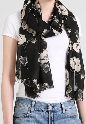Écharpe noire à motif floral beige drapée autour du cou, portée sur une chemise blanche à manches courtes et un jean en denim bleu.