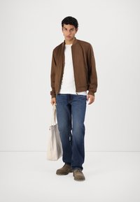 Brun bomberjakke med en struktureret overflade, hvid T-shirt, blå jeans og brune sko. Holder en let kanvas tote bag.