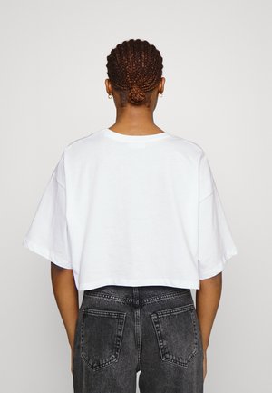 Personne avec les cheveux tressés en chignon portant un t-shirt blanc ample et court et un jean noir délavé taille haute, vue de dos.