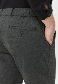 Mann in dunkelgrauen Hosen mit einer geknöpften Gesäßtasche und einer Hand im vorderen Taschen, aus der Hüfte nach unten gezeigt.