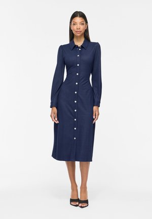 Femme debout portant une robe midi bleu marine à manches longues, boutonnée, avec un col et des talons noirs ouverts à l’avant.