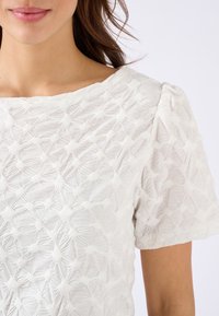 Witte textuur blouse met een bloemenachtig patroon, bootnek en korte pofmouwen. Zachte stof met subtiele verhoogde accenten.