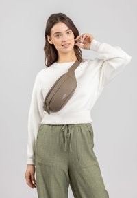 Bruine leren crossbody tas met een gebogen vorm, dubbele ritssluiting en verstelbare schouderriem, gedragen over een witte trui en groene broek.
