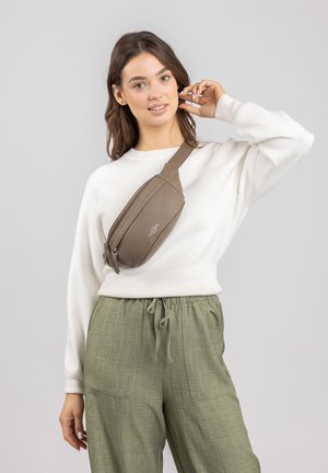 Brown leather crossbody bag with a curved shape, double zip closure, and adjustable strap, worn over a white sweater and green trousers.