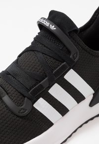 Svart atletisk sko med texturerad mesh-ovandel, svarta snören och tre vita ränder. Har en svart loggtagg och en vadderad vit sula.