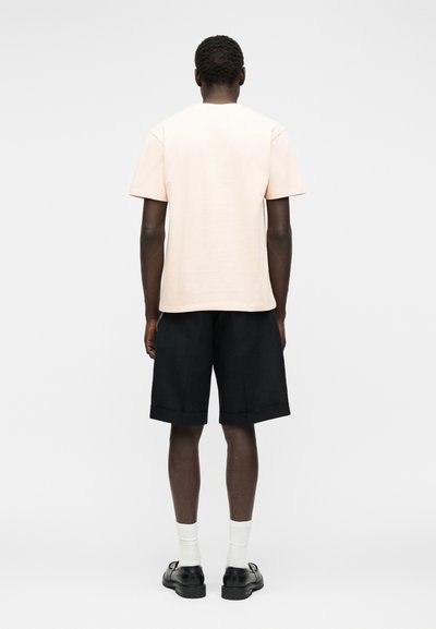 Άνδρας που στέκεται με την πλάτη, φοράει ανοιχτό μπεζ κοντομάνικο πουκάμισο, μαύρα σορτς μέχρι το γόνατο, λευκές κάλτσες και μαύρα παπούτσια.