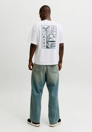 Mann trägt weißes T-Shirt mit schwarzem japanischem Landschafts- und Blumenprint auf dem Rücken, kombiniert mit ausgewaschenen blauen Jeans und schwarzen Sneakern.