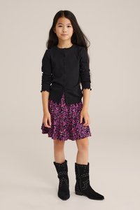 Černý kardigan s dlouhými rukávy a knoflíky, kombinovaný s fialovou květinovou sukní s volánky a černými kotníkovými botami se cvočky. Jednoduché pozadí.