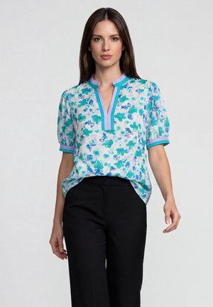 Jonge vrouw die een korte mouwen bloemige blouse met turquoise en paarse accenten en een zwarte broek draagt, staand tegen een effen lichte achtergrond.
