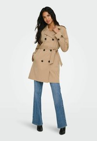 Femme portant un trench-coat beige à double boutonnage avec ceinture, jean évasé bleu et bottines noires, posant avec une main dans la poche.